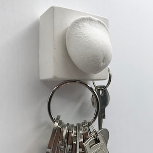 Puede incluir: Porta llaves de cerámica blanca con un diseño redondeado y texturado. Un juego de llaves está unido al porta llaves.