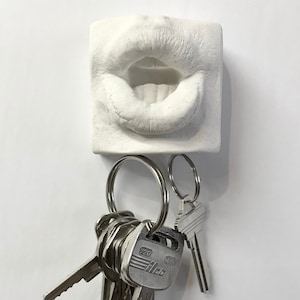 Könnte beinhalten: Ein weißer, quadratischer Wand-Schlüsselhalter mit einem realistischen, skulpturalen Relief von geöffneten menschlichen Lippen. Das Objekt verfügt über zwei silberfarbene Metallringe an der Unterseite, an denen mehrere silberne Metallschlüssel hängen.