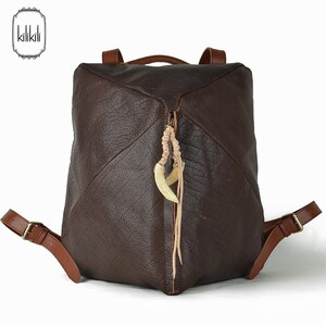 May include: A brown leather backpack with a unique triangular design. The backpack has a zipper closure and a decorative bone charm hanging from the zipper pull.
