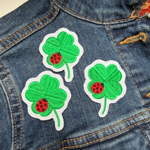 May include: Three embroidered patches of green four-leaf clovers with red ladybirds, each with a white border. They are displayed on a blue denim jacket.