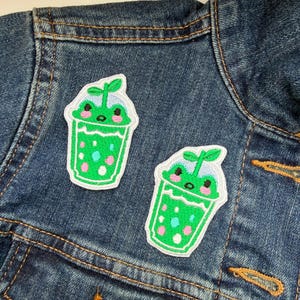 May include: Two embroidered patches depicting a green cartoon drink with a smiling frog face and a sprout. The patches have a white border and are attached to a denim jacket.