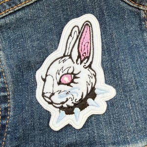 May include: Embroidered patch of a white rabbit head with pink inner ears and a black collar with light blue spikes. The rabbit has pink eyes, set against a blue denim background. The patch is attached to a denim jacket.