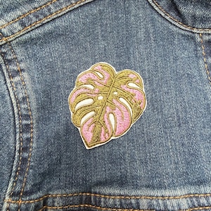 May include: A colourful embroidered patch depicting a monstera leaf in pink and green. The patch is attached to a blue denim jacket, highlighting the detailed stitching and bright colours. This decorative patch adds a touch of nature-inspired style to clothing and accessories.