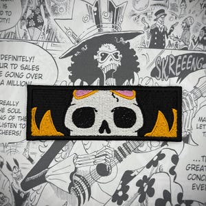 May include: Black and white embroidered patch featuring a skull and crossbones design. The skull is white with black eyes and a pink nose. The crossbones are yellow. The patch is edged with a black border.