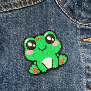 May include: A green embroidered frog patch with black eyes, pink cheeks, and a white belly is sewn onto a denim jacket. The frog has a smiling expression and is a cute, whimsical design.