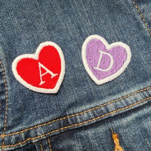 May include: Two embroidered heart patches with white borders and letters on a denim jacket. One heart is red with a white "A", the other is purple with a white "D". The denim jacket has visible stitching and a button.
