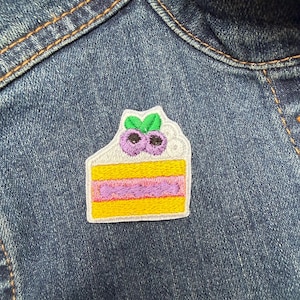 May include: Embroidered patch of a slice of cake with yellow, purple, and white details, topped with blueberries and green leaves. The patch is sewn onto a blue denim jacket.