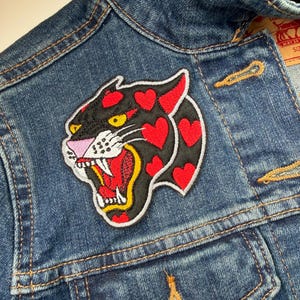 May include: A close-up of a denim jacket with a decorative patch. The patch depicts a black panther head with red heart-shaped spots, a pink nose, and a red open mouth. The jacket has a button closure and pockets.