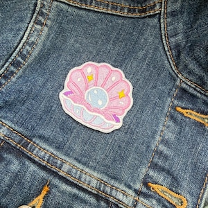 May include: A pink, blue, and yellow embroidered seashell patch is attached to a blue denim jacket. The patch features a pearl and star accents. The jacket has orange stitching and a silver button.