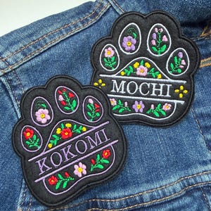 May include: Two black paw-shaped embroidered patches with floral designs and names. One patch says "MOCHI" in white, the other says "KOKOMI" in white. The patches are decorated with colourful flowers and green leaves, set against a blue denim background.