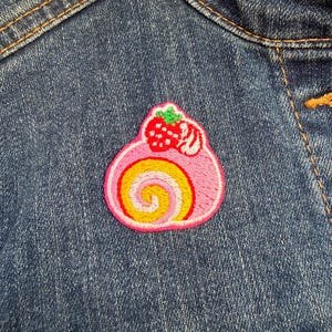 May include: A pink embroidered patch of a Swiss roll cake with a strawberry on top. The patch features a yellow and white swirl centre and is sewn onto a blue denim jacket.