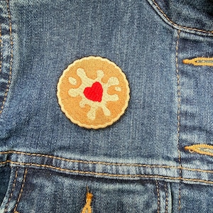 May include: A close-up of a denim jacket with a biscuit-shaped patch. The patch is tan with a red heart in the middle. The jacket has gold stitching and a buttoned pocket.