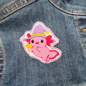 May include: A pink axolotl patch wearing a purple witch's hat with yellow details and stars. The patch is outlined in white and is on a blue denim jacket.