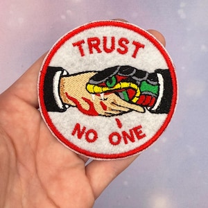 May include: Circular embroidered patch with the text "TRUST NO ONE" in red. The design features a handshake with a snake coiled around it. The snake is green, yellow, and red. The patch has a white background and a red border.