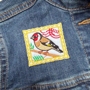 May include: A denim jacket with a square patch showing an embroidered bird design. The patch has a yellow border and depicts a bird perched on a branch, with red, brown, and black details. The jacket has visible stitching.