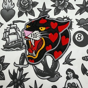 May include: Embroidered patch of a black panther with red heart-shaped spots, open mouth, and yellow fangs. The patch is surrounded by traditional tattoo-style illustrations in black and grey, including roses, stars, and a ship.