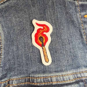 May include: Embroidered patch of a burning match, with red, orange, and yellow flames. The matchstick has a brown wooden handle and a black head. The patch is outlined in white and is attached to a blue denim jacket.