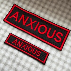 May include: Two rectangular embroidered patches featuring a black background with a red border and the word ANXIOUS stitched in red capital letters. The patches are displayed in two different sizes, both with a textured fabric finish.