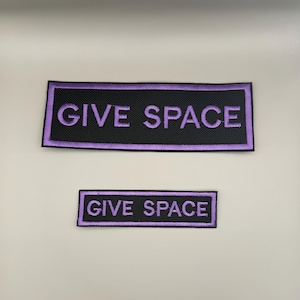 May include: Two rectangular embroidered patches featuring a black background with a lavender border and the text GIVE SPACE in lavender capital letters. The patches are identical in design, with one patch significantly larger than the other.