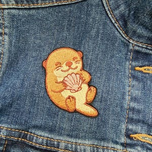 May include: Embroidered patch of a smiling otter holding a seashell, in shades of tan, orange, and cream. The patch is sewn onto a blue denim jacket. The otter patch is a cute, whimsical design.