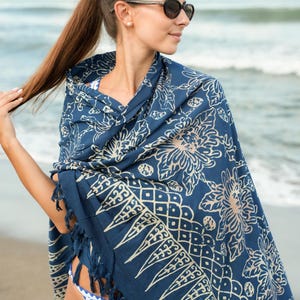Può includere: Uno scialle blu navy con motivo floreale beige e frange. Lo scialle è drappeggiato sulle spalle. La persona indossa occhiali da sole e un bikini. Lo sfondo è una spiaggia con onde.