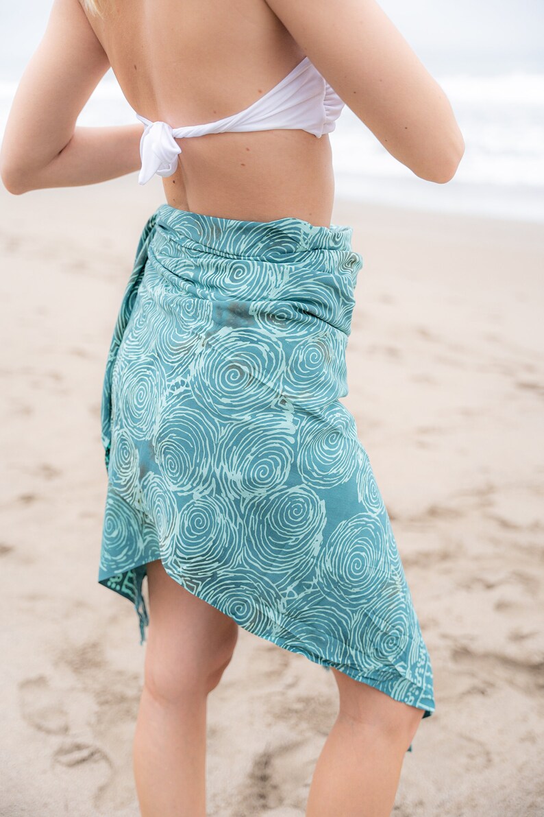 May include: Teal sarong with a white spiral pattern, tied at the waist. The sarong is worn over a white bikini top. The image is taken on a sandy beach, with the ocean visible in the background.