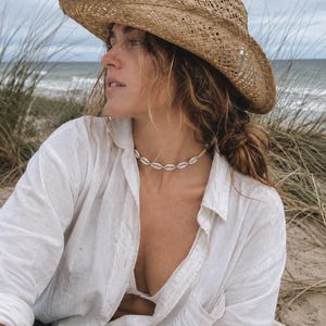 May include: A tan woven straw hat with a wide brim and a white cowrie shell choker necklace. The necklace features a series of small, off-white shells strung together in a circular pattern, resting against the neck.
