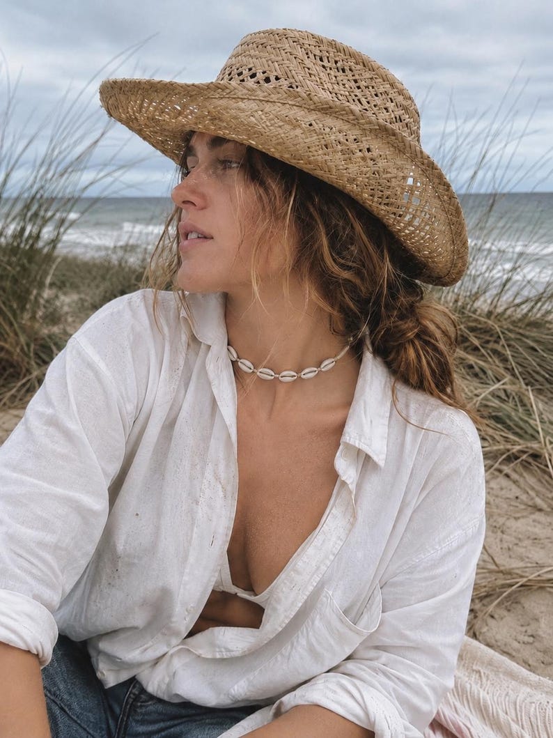 May include: A tan woven straw hat with a wide brim and a white cowrie shell choker necklace. The necklace features a series of small, off-white shells strung together in a circular pattern, resting against the neck.