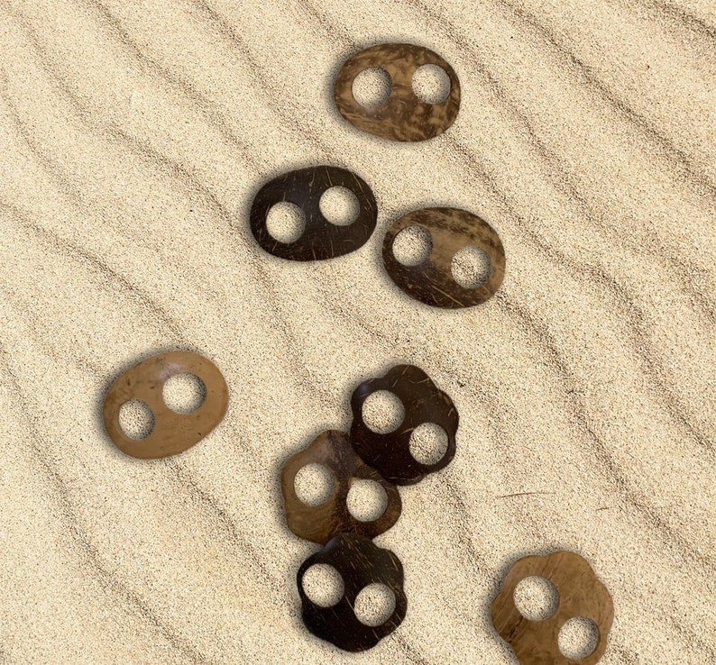 May include: A collection of oval-shaped, natural-toned coconut shell buttons with two holes each. The buttons vary in color from light tan to dark brown, scattered on a sandy surface. These buttons are ideal for sewing and craft projects.