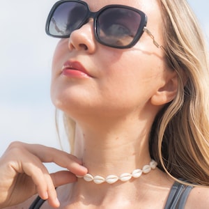 May include: A choker necklace made of small, white, oval-shaped cowrie shells strung together in a continuous row, worn around the neck. The necklace features a simple design with the natural, off-white texture of the shells clearly visible.