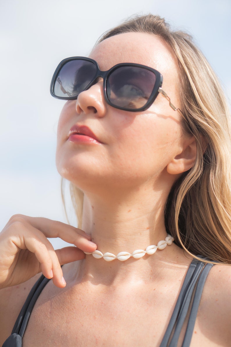 May include: A choker necklace made of small, white, oval-shaped cowrie shells strung together in a continuous row, worn around the neck. The necklace features a simple design with the natural, off-white texture of the shells clearly visible.