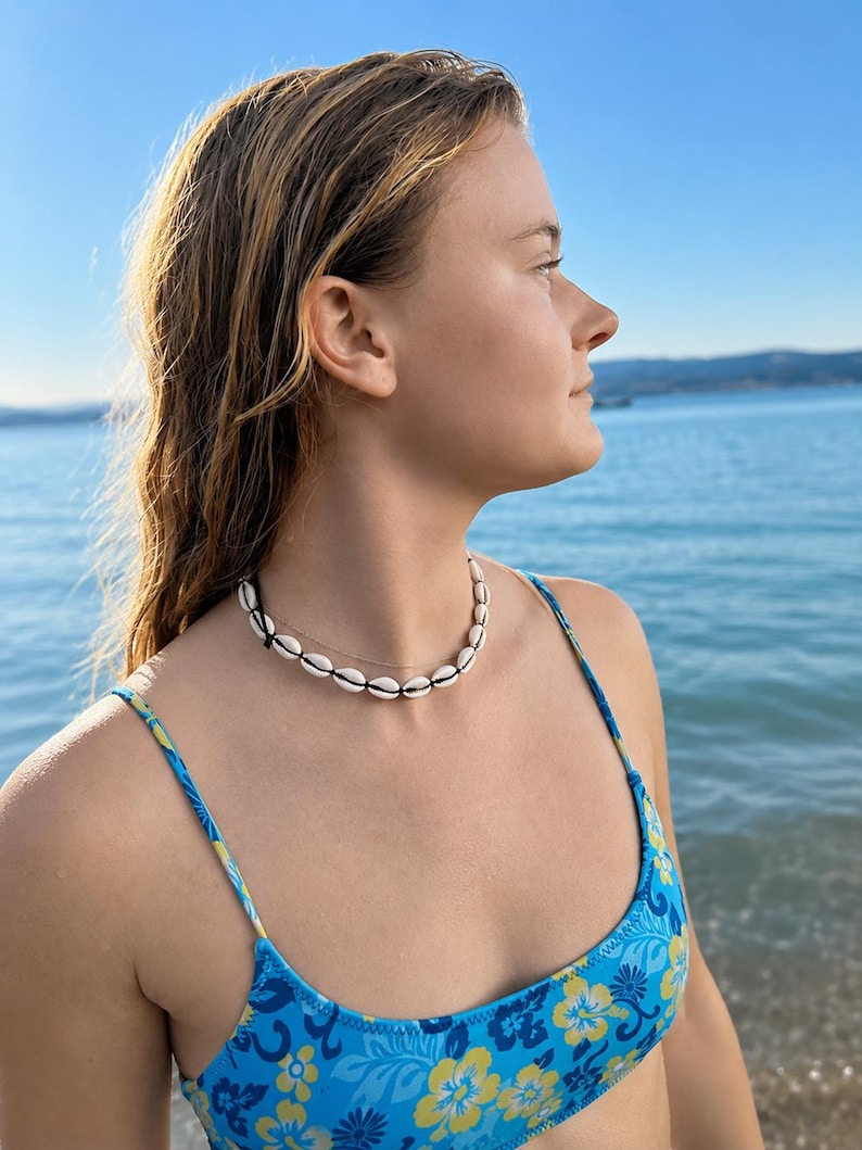 May include: A choker necklace featuring a single strand of white cowrie shells strung on a thin black cord, worn alongside a delicate, thin metallic chain necklace. The jewellery is displayed against a bright blue floral patterned bikini top.