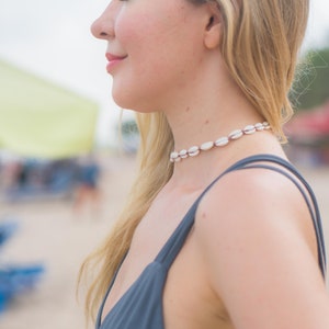 May include: A choker necklace featuring a series of small, white, oval-shaped cowrie shells strung together on a dark cord. The necklace sits closely around the neck, showcasing the natural texture and off-white colour of the individual shells.