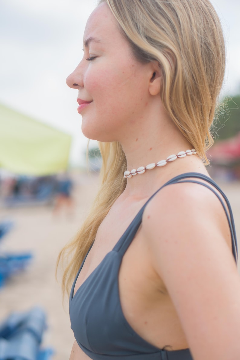 May include: A choker necklace featuring a series of small, white, oval-shaped cowrie shells strung together on a dark cord. The necklace sits closely around the neck, showcasing the natural texture and off-white colour of the individual shells.