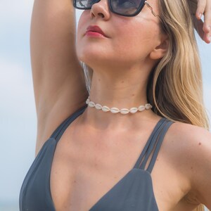 May include: A white cowrie shell choker necklace worn with a dark grey bikini top featuring double straps and black oversized sunglasses with dark lenses. The necklace consists of a single row of small, uniform, off-white sea shells strung together.