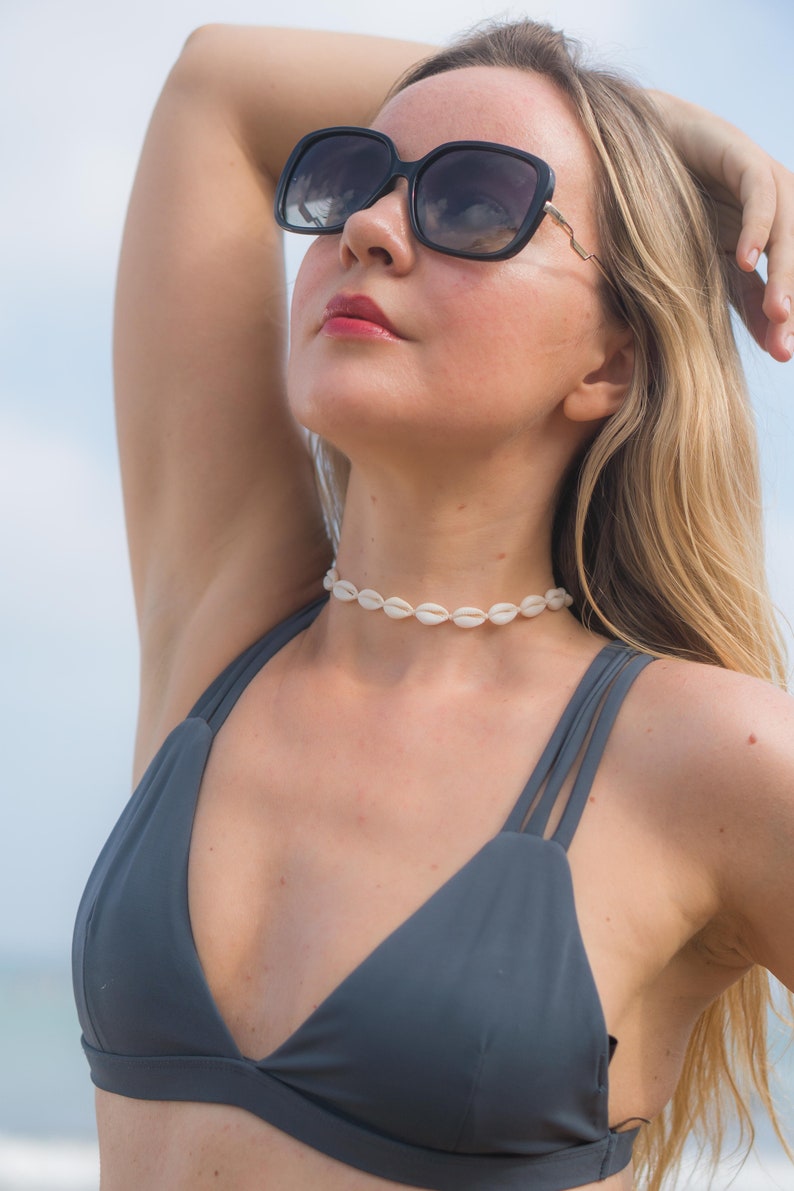 May include: A white cowrie shell choker necklace worn with a dark grey bikini top featuring double straps and black oversized sunglasses with dark lenses. The necklace consists of a single row of small, uniform, off-white sea shells strung together.