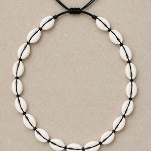May include: A necklace featuring a series of white cowrie shells strung along a thin black cord. The cord is arranged in a circular shape and includes an adjustable sliding knot mechanism at the top, allowing for variations in the length of the necklace.