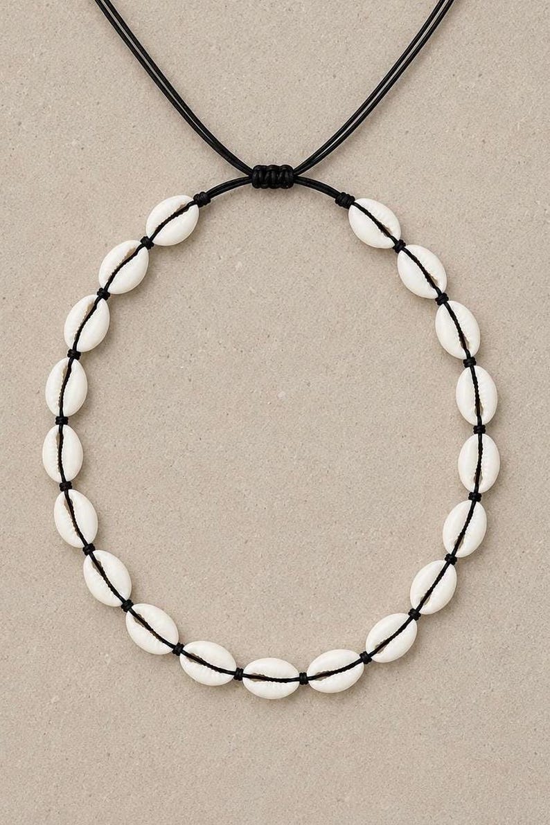 May include: A necklace featuring a series of white cowrie shells strung along a thin black cord. The cord is arranged in a circular shape and includes an adjustable sliding knot mechanism at the top, allowing for variations in the length of the necklace.
