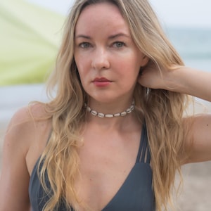 May include: A dark grey bikini top featuring a V-neckline and triple spaghetti straps on each shoulder. The garment is paired with a choker necklace made of small, white, oval-shaped seashells strung together in a single row around the neck.
