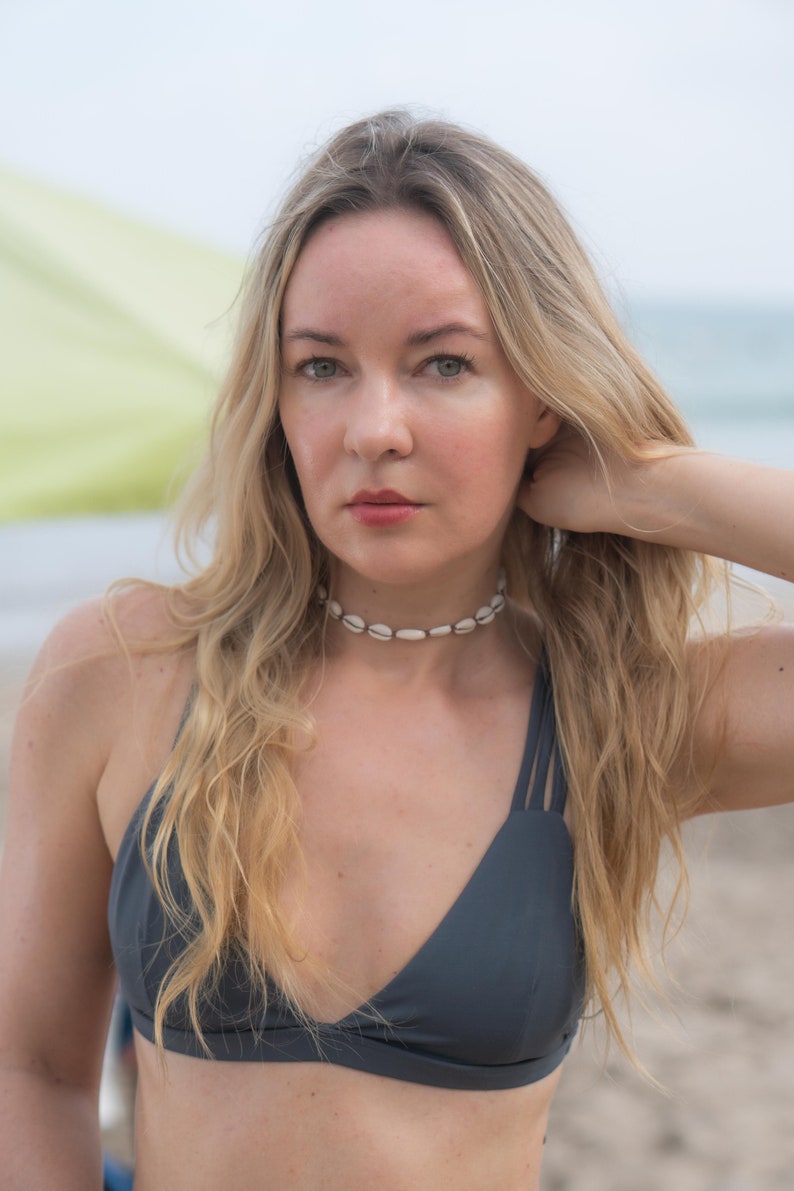 May include: A dark grey bikini top featuring a V-neckline and triple spaghetti straps on each shoulder. The garment is paired with a choker necklace made of small, white, oval-shaped seashells strung together in a single row around the neck.