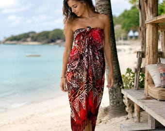 May include: A strapless sarong wrap dress featuring a vibrant red, black, and white batik-style pattern with intricate floral and geometric motifs. The garment is tied at the chest and finished with a fringed hemline that falls below the knees.