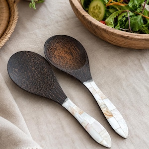 May include: Two dark brown wooden serving spoons with rounded bowls and handles inlaid with a mosaic of iridescent white and cream-coloured shell tiles. The spoons feature a visible natural wood grain texture and a smooth, polished finish on the handles.