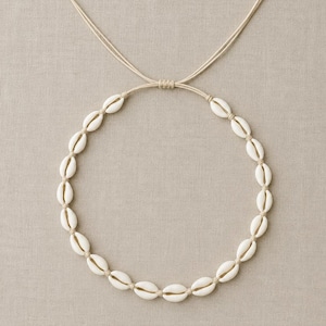 May include: A necklace featuring a circular arrangement of white cowrie shells strung on a light beige cord. The cord includes an adjustable sliding knot mechanism at the top, allowing for variations in length to accommodate different neck sizes.