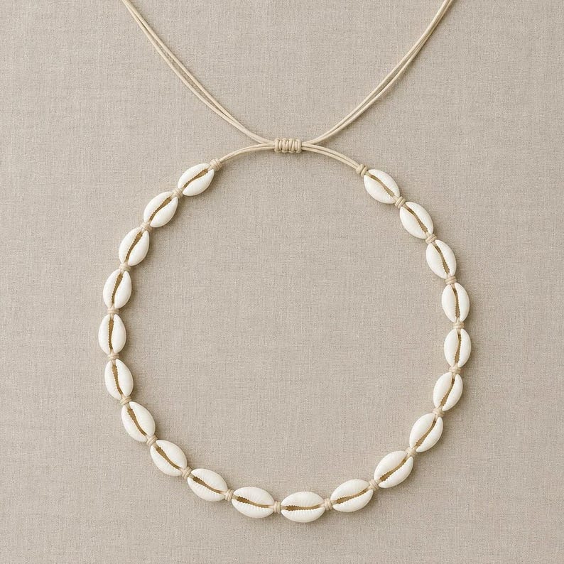 May include: A necklace featuring a circular arrangement of white cowrie shells strung on a light beige cord. The cord includes an adjustable sliding knot mechanism at the top, allowing for variations in length to accommodate different neck sizes.