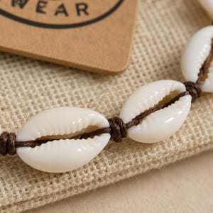 May include: A bracelet featuring white cowrie shells strung on a dark brown cord. Each shell is separated by a small, tight knot in the cord. The shells are smooth, oval-shaped, and have a central opening with ribbed edges along the length.