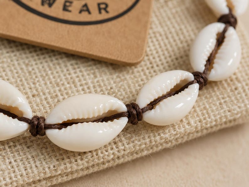 May include: A bracelet featuring white cowrie shells strung on a dark brown cord. Each shell is separated by a small, tight knot in the cord. The shells are smooth, oval-shaped, and have a central opening with ribbed edges along the length.