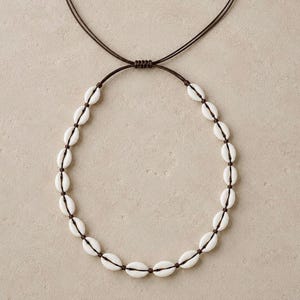 May include: A necklace featuring a series of white cowrie shells strung along a dark brown cord. The design includes an adjustable sliding knot mechanism at the top, allowing the length of the cord to be modified for a comfortable fit.
