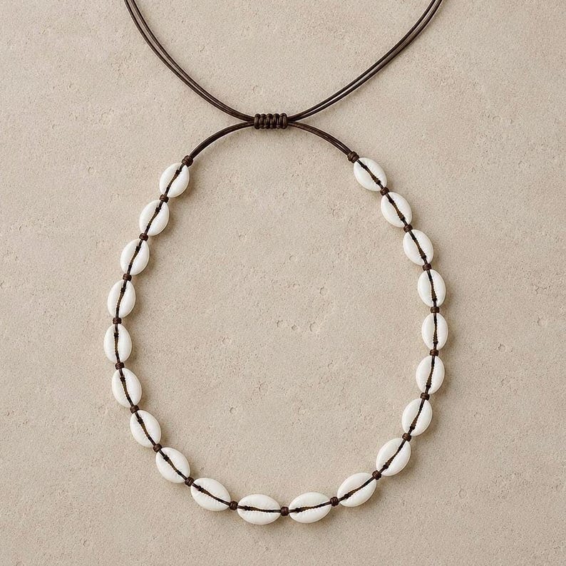 May include: A necklace featuring a series of white cowrie shells strung along a dark brown cord. The design includes an adjustable sliding knot mechanism at the top, allowing the length of the cord to be modified for a comfortable fit.
