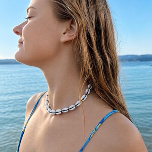 May include: A choker necklace featuring a single strand of white cowrie shells strung together on a thin black cord. The shells are arranged in a continuous circular pattern, creating a simple and uniform design that rests around the neck.