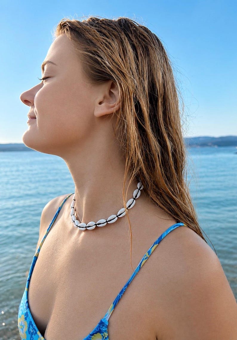 May include: A choker necklace featuring a single strand of white cowrie shells strung together on a thin black cord. The shells are arranged in a continuous circular pattern, creating a simple and uniform design that rests around the neck.